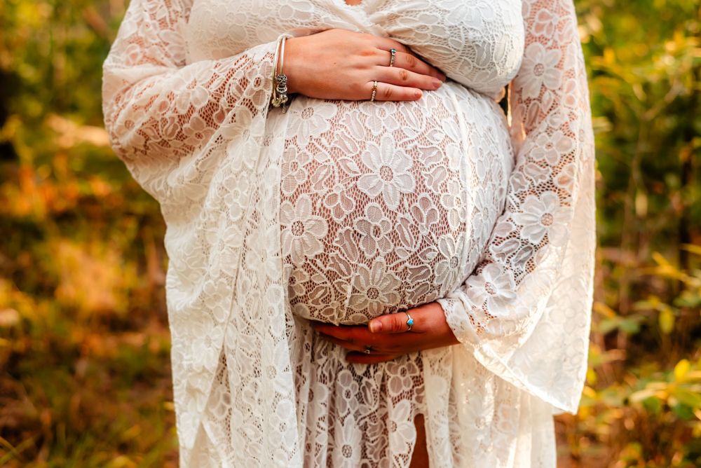 Hands rest on a white lace maternity dress against an autumn backdrop.