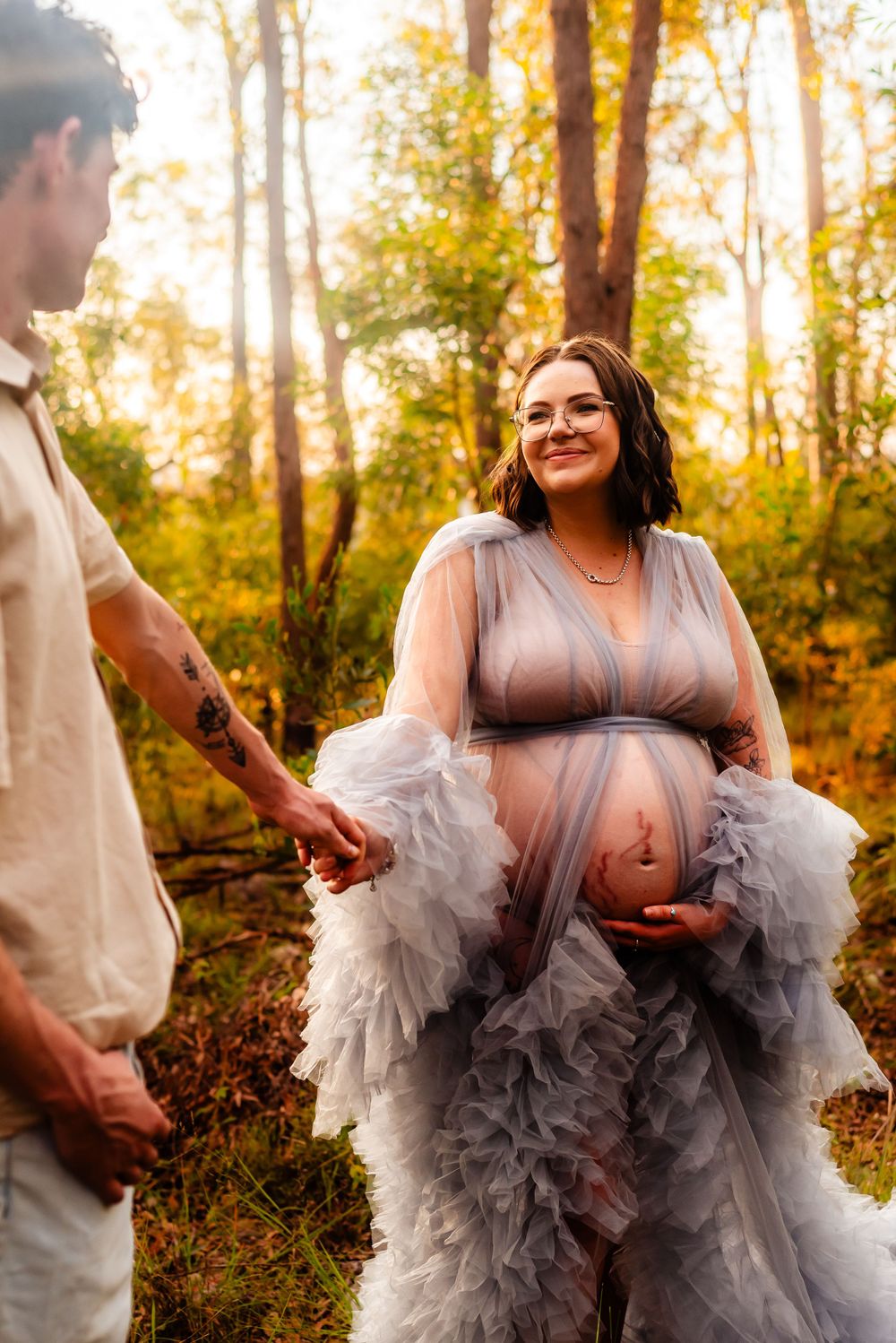 Expectant couple sharing loving moment in ethereal forest maternity shoot with flowing gray tulle dress at sunset.