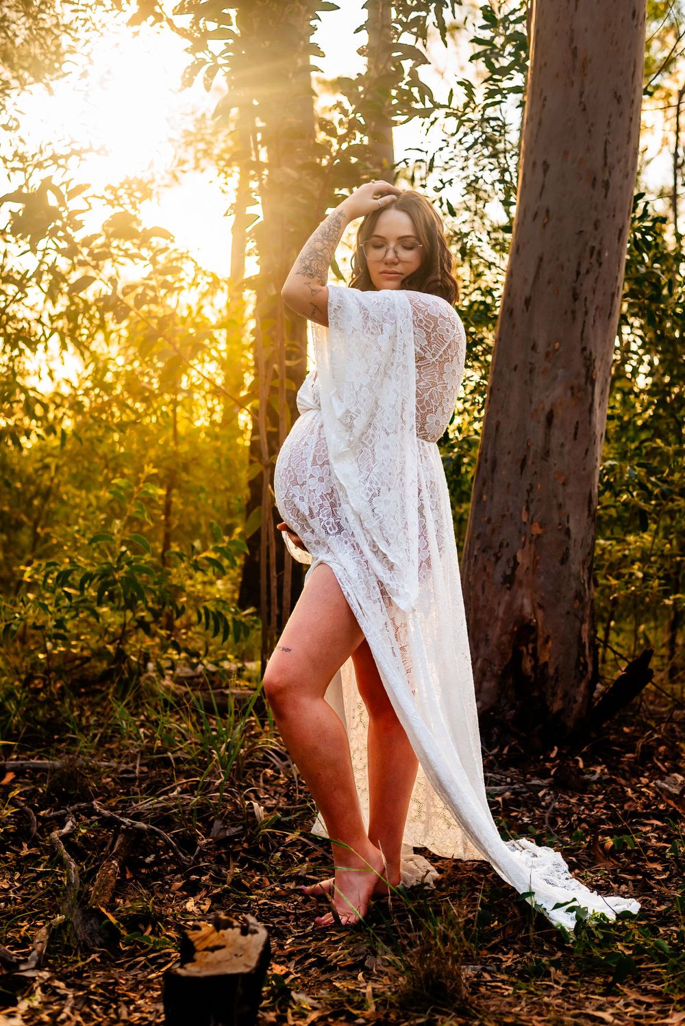 Maternity photo of woman in white lace dress posing elegantly in forest with sunlight filtering through trees.