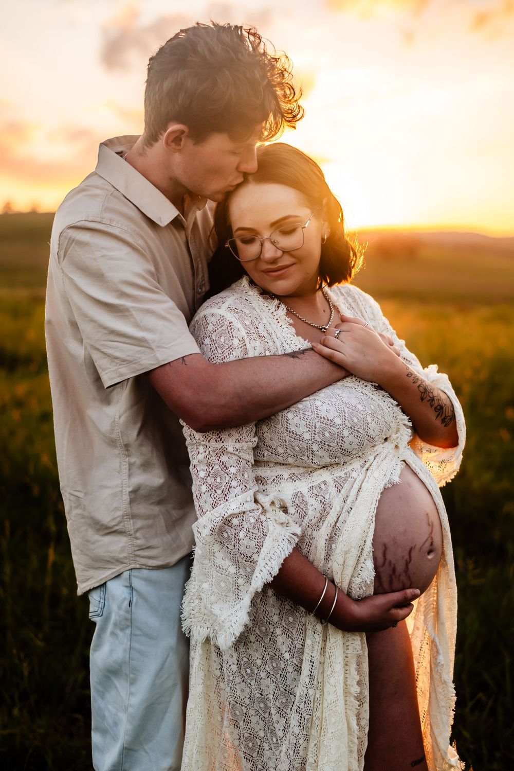 Intimate maternity portrait of couple embracing at sunset in white lace dress.
