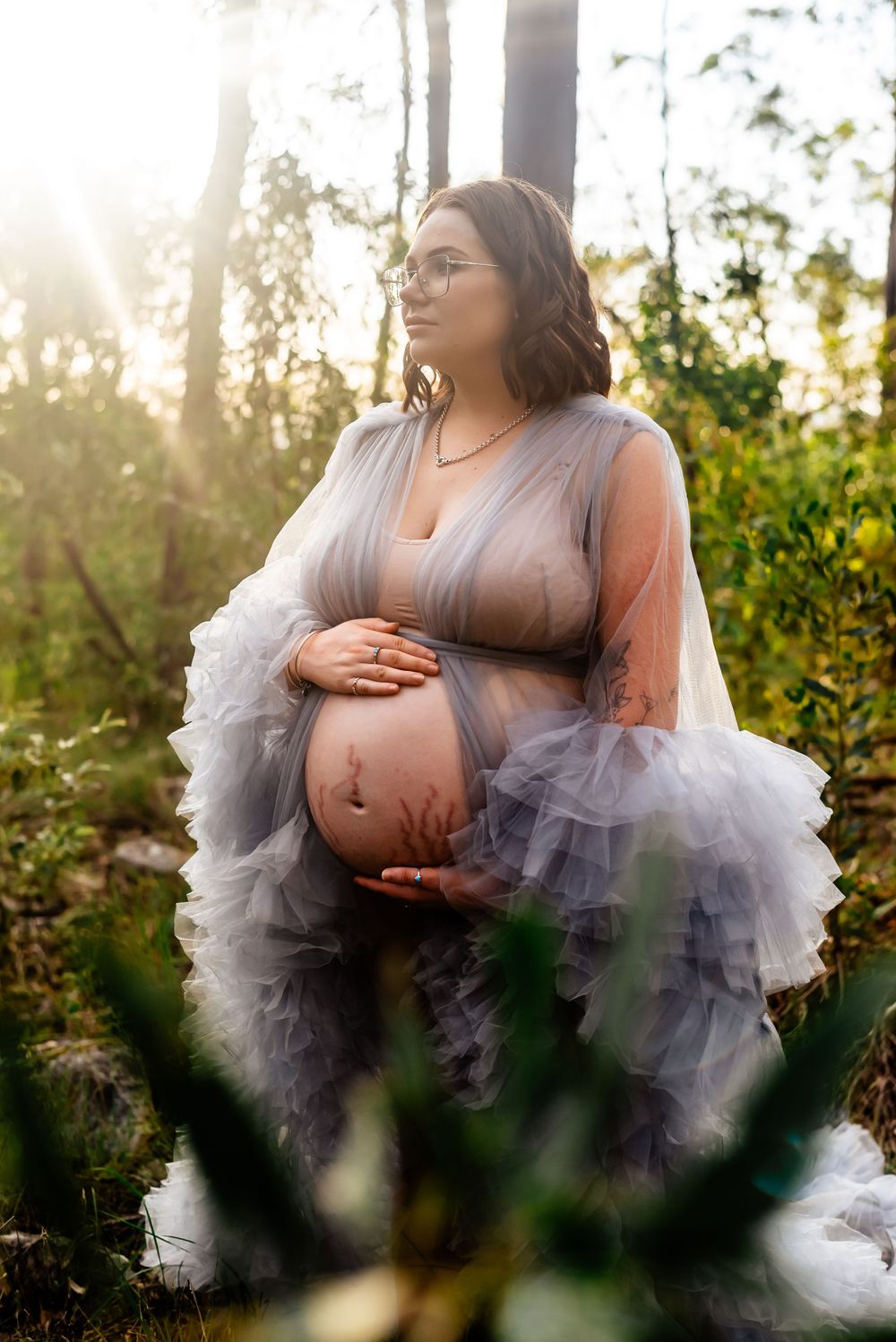 Pregnant woman in lavender tulle dress poses in sunlit forest setting.