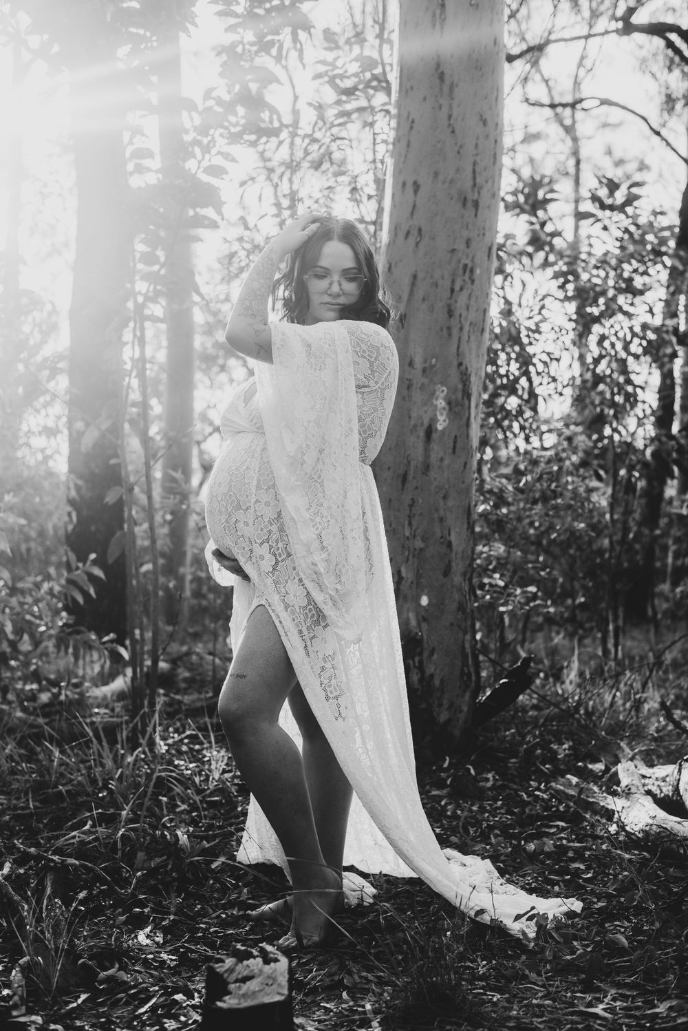Artistic black and white maternity photo of woman in lace wrap dress in forest setting.