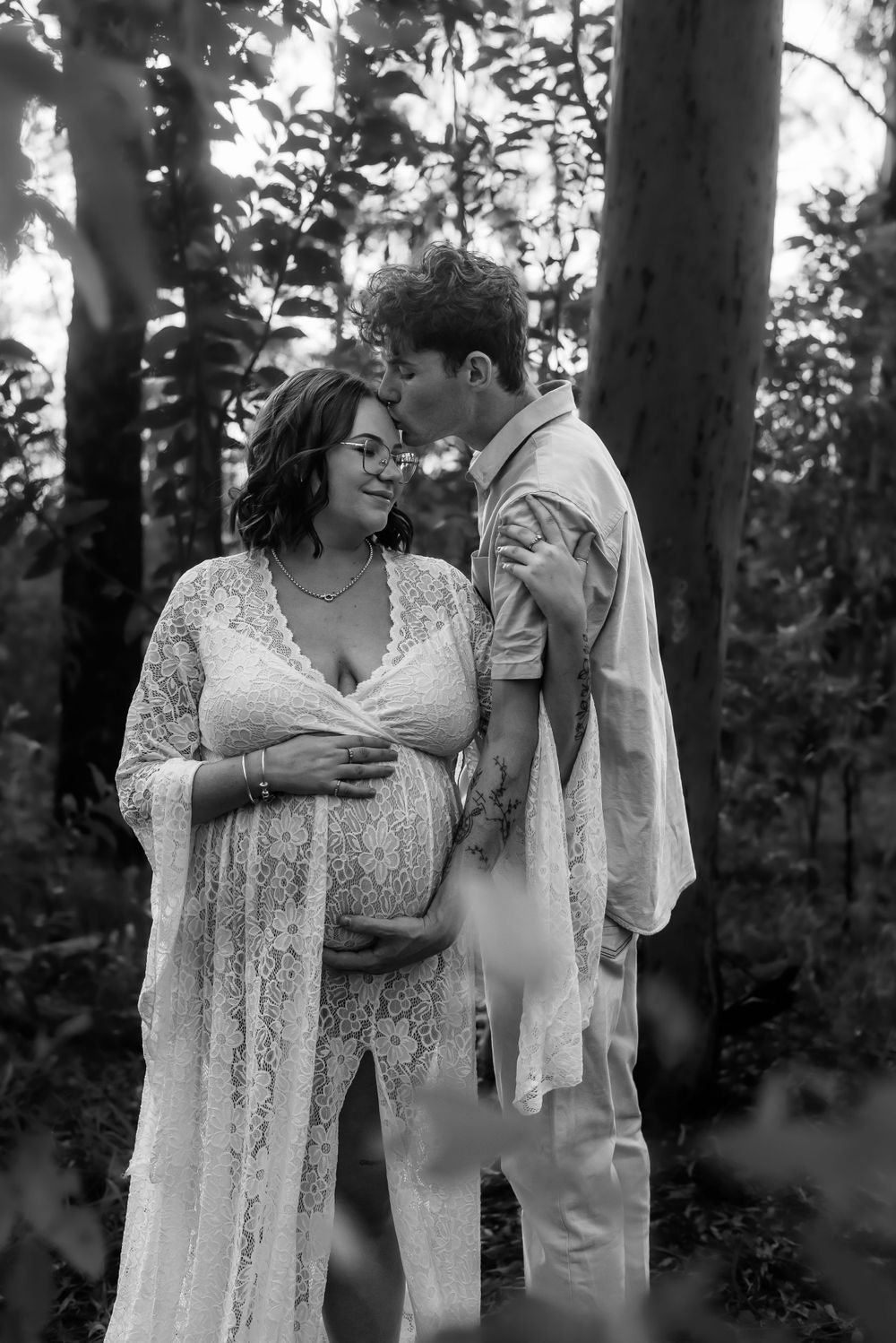 Black and white maternity portrait of couple sharing tender moment among trees.