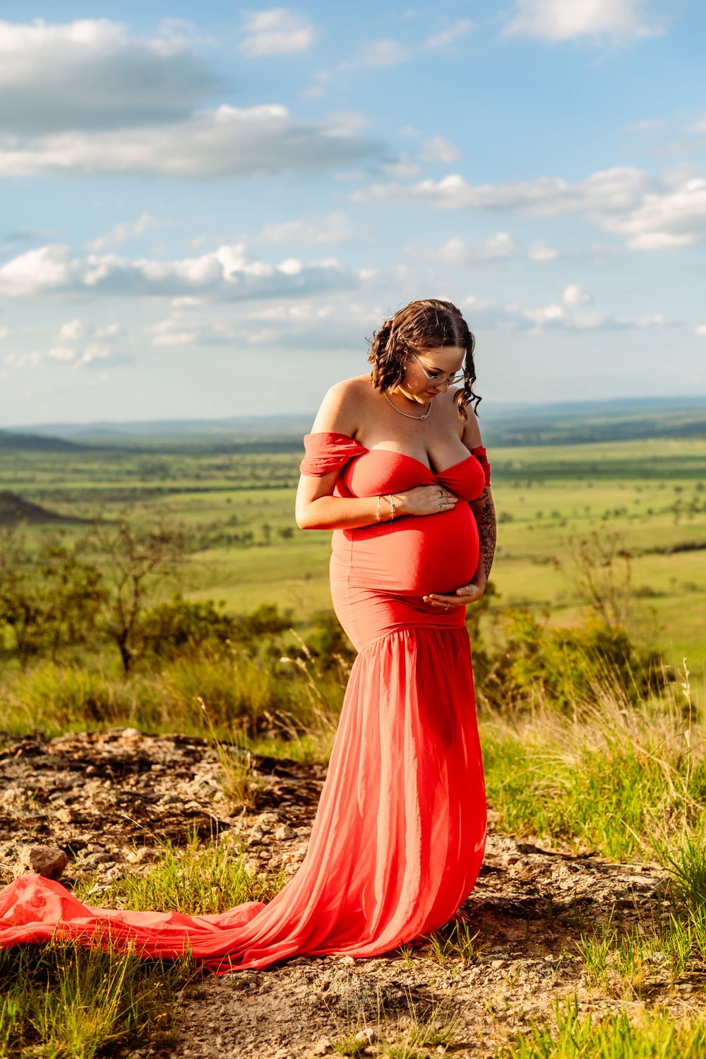 Pregnant woman in flowing red dress poses on hilltop against expansive grassland vista.