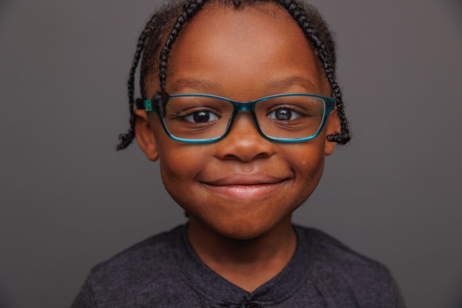 Portrait of a child with braided hair and glasses wearing a dark top against gray background.