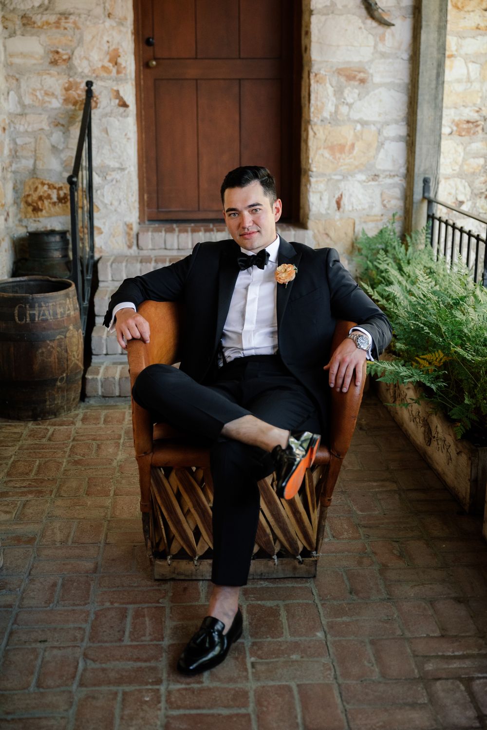 A groom in a black tuxedo relaxes in a leather chair on a stone porch.
