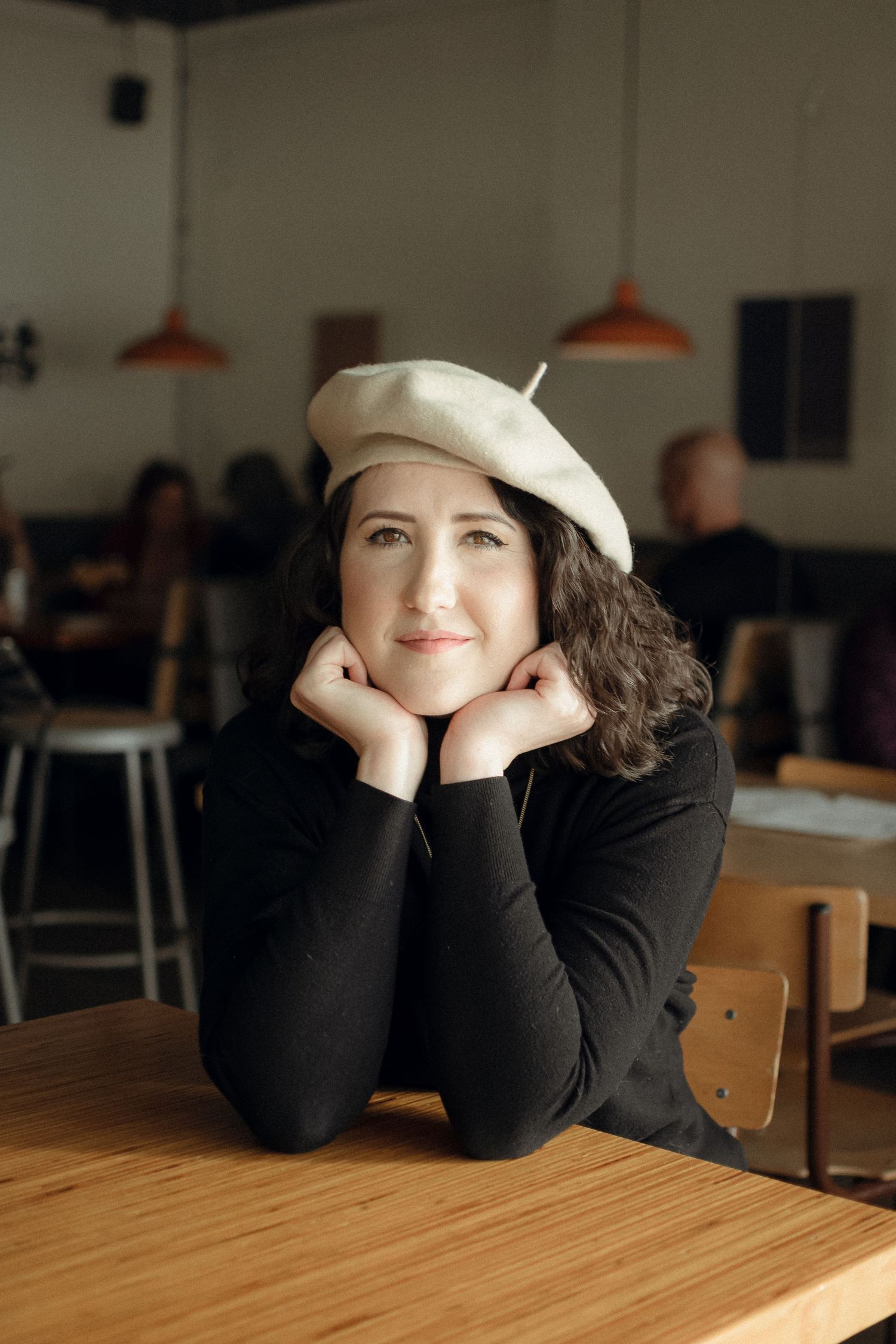 Someone in a white beret and black sweater rests their head on their hands at a cafe table.