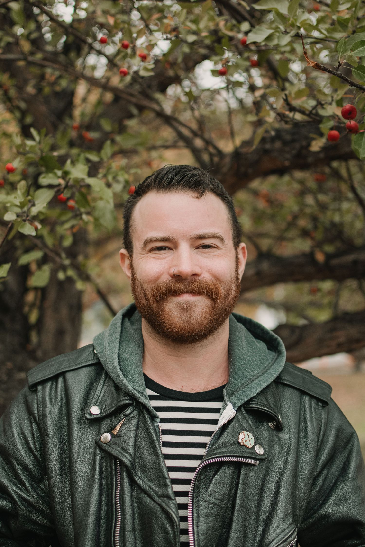 A person in striped shirt and green jacket poses in front of an apple tree.