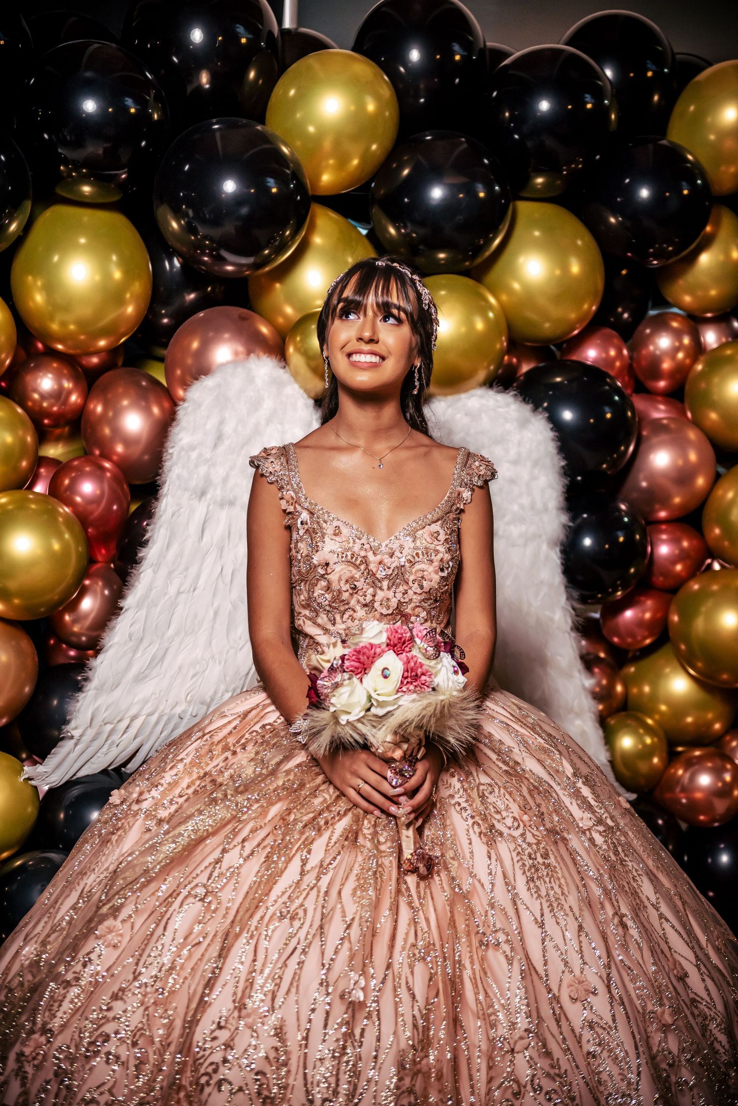 Rose gold quinceañera dress with angel wings against black and gold balloon backdrop.