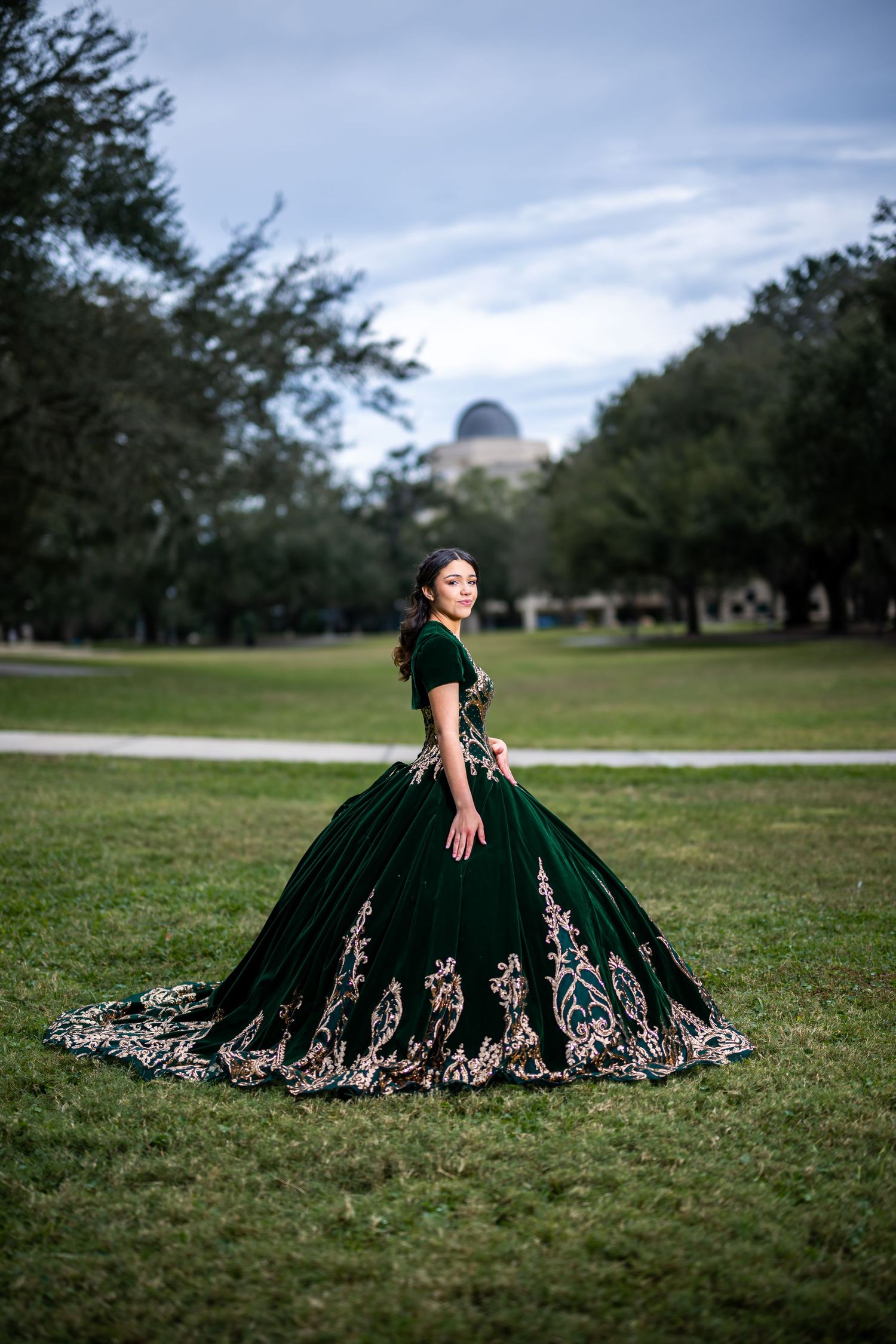 Dark green velvet ballgown with gold detailing photographed on a grassy lawn with trees.