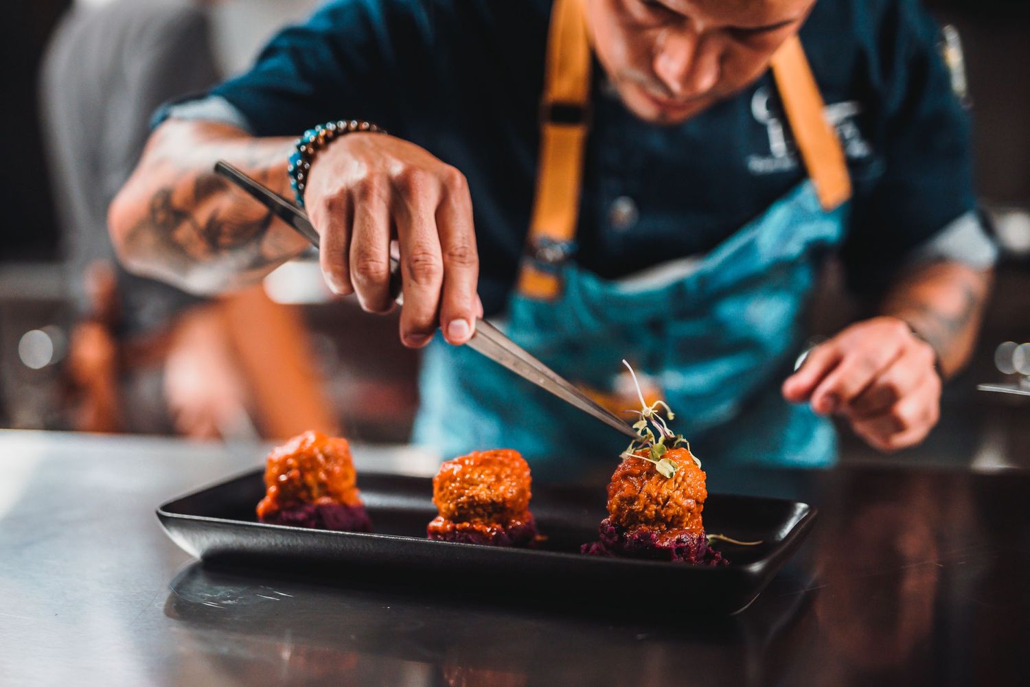 Chef plating golden brown croquettes on black serving plate with flame in background.