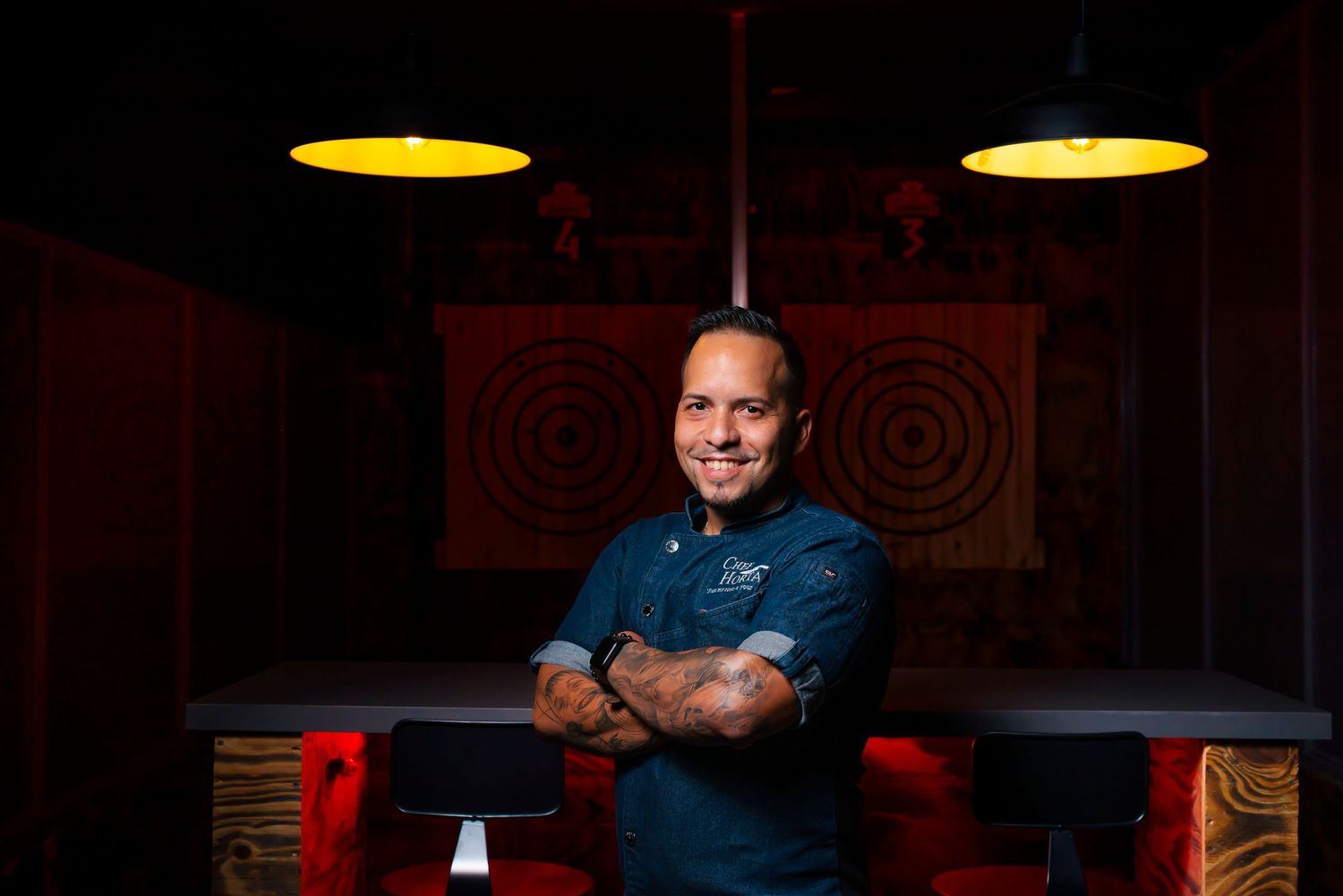 Professional chef standing in restaurant kitchen with dramatic lighting and tattoos.