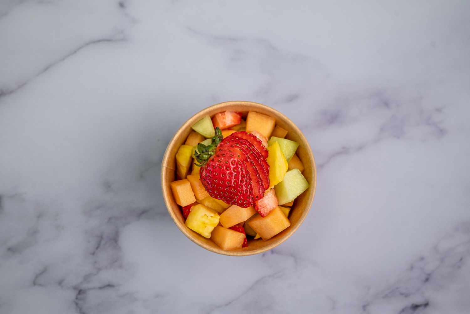 Fresh fruit bowl with diced tropical fruits and a large strawberry garnish on marble surface.