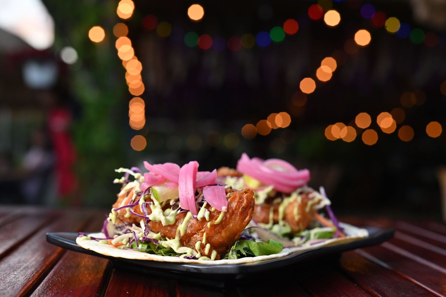 Colorful tacos garnished with pink sauce and fresh herbs against bokeh lights background.