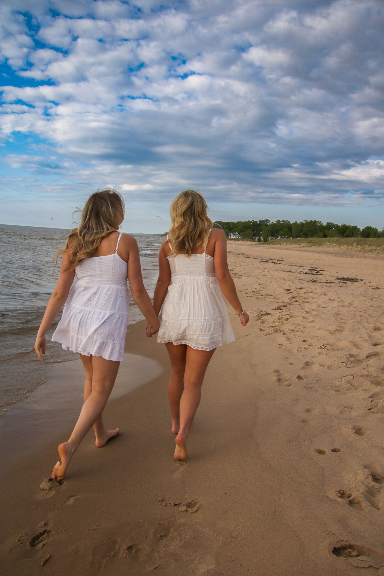 Two people in white dresses walking and leaving footprints along a sandy beach.