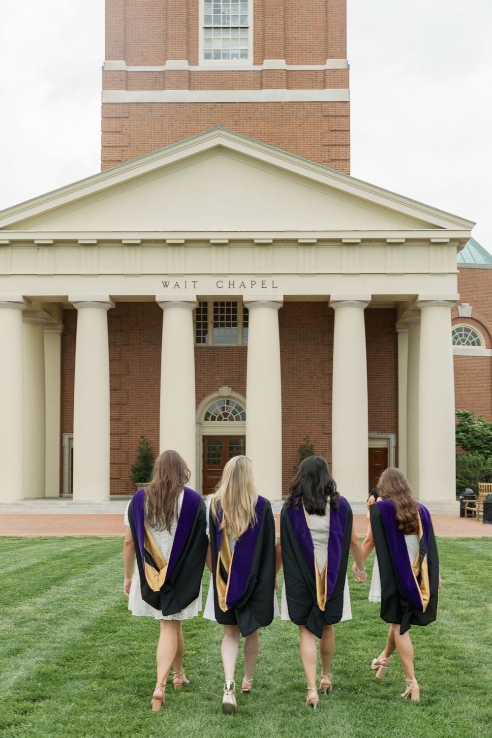 Four graduates in purple and gold stoles photographed from behind in front of Wait Chapel.