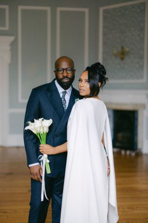 Elegant couple in wedding attire pose while holding white calla lilies in a classic indoor setting.