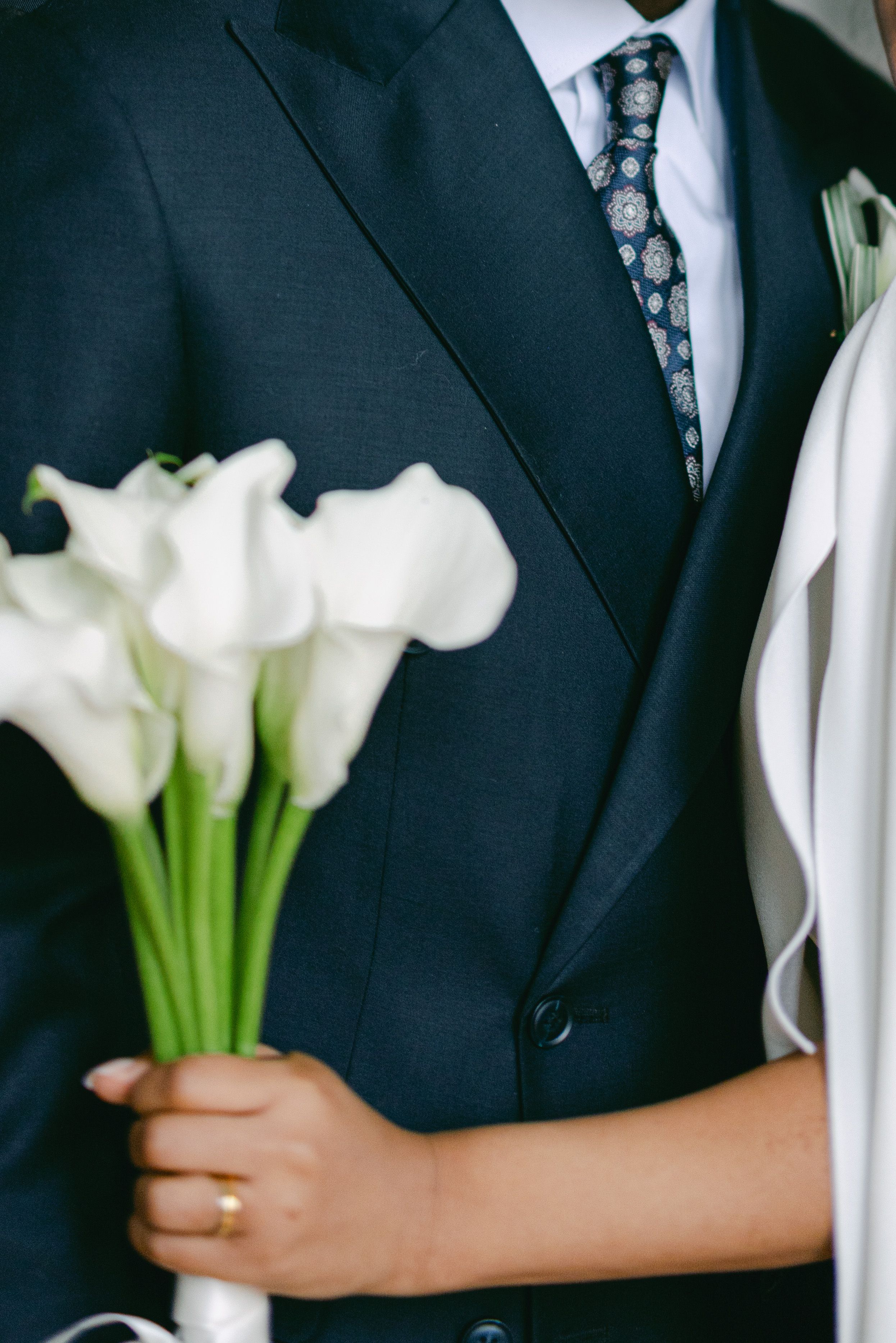Close-up of white calla lily bouquet held against navy blue suit with patterned tie.