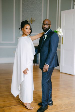 Modern wedding couple poses in stylish navy suit and flowing white cape dress against grey walls.