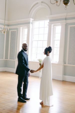 Romantic wedding couple holds hands in sunlit room with classic architectural details.