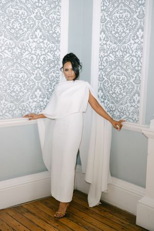 Bride in modern white cape dress poses against damask wallpaper in elegant room.