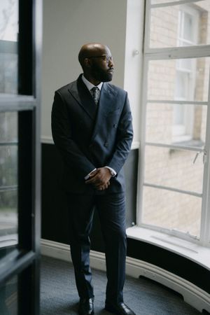 Professional in a dark suit stands contemplatively by a window.