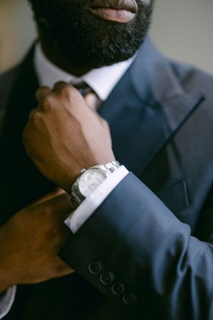 Close-up of a hand adjusting a necktie while wearing a silver watch and navy suit.