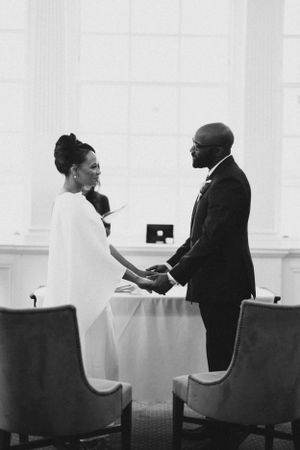 Intimate black and white wedding ceremony moment showing couple holding hands in an elegant indoor setting.