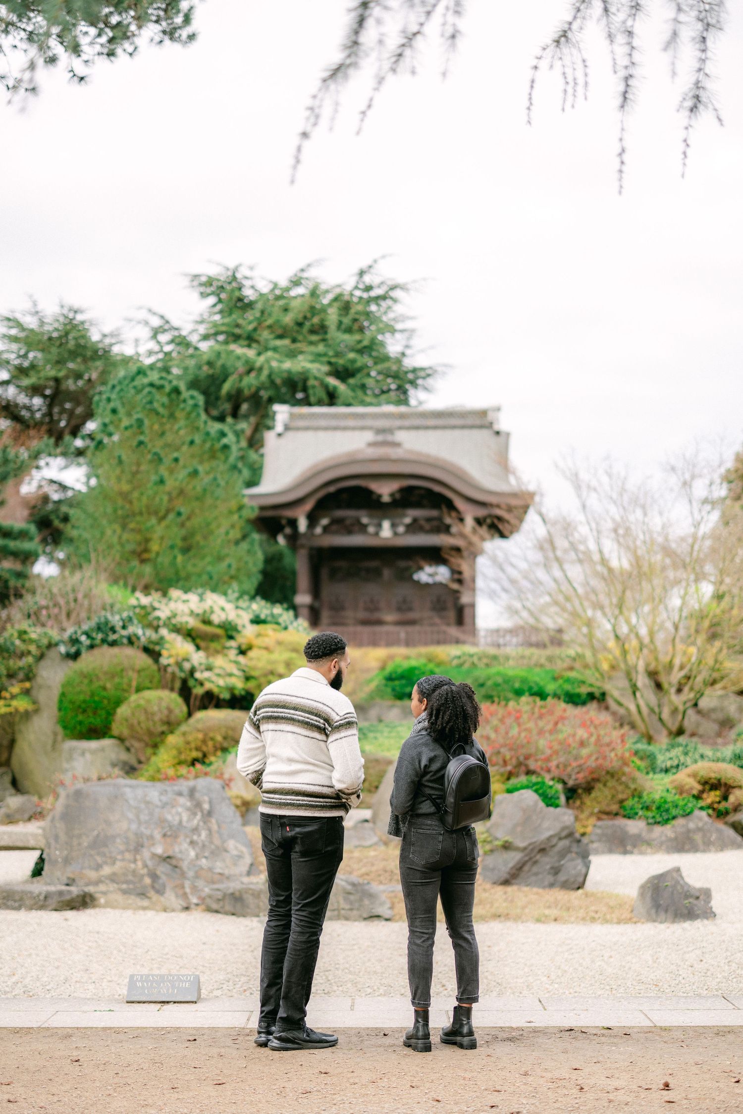 Two people admiring a traditional Japanese garden pavilion surrounded by manicured shrubs.