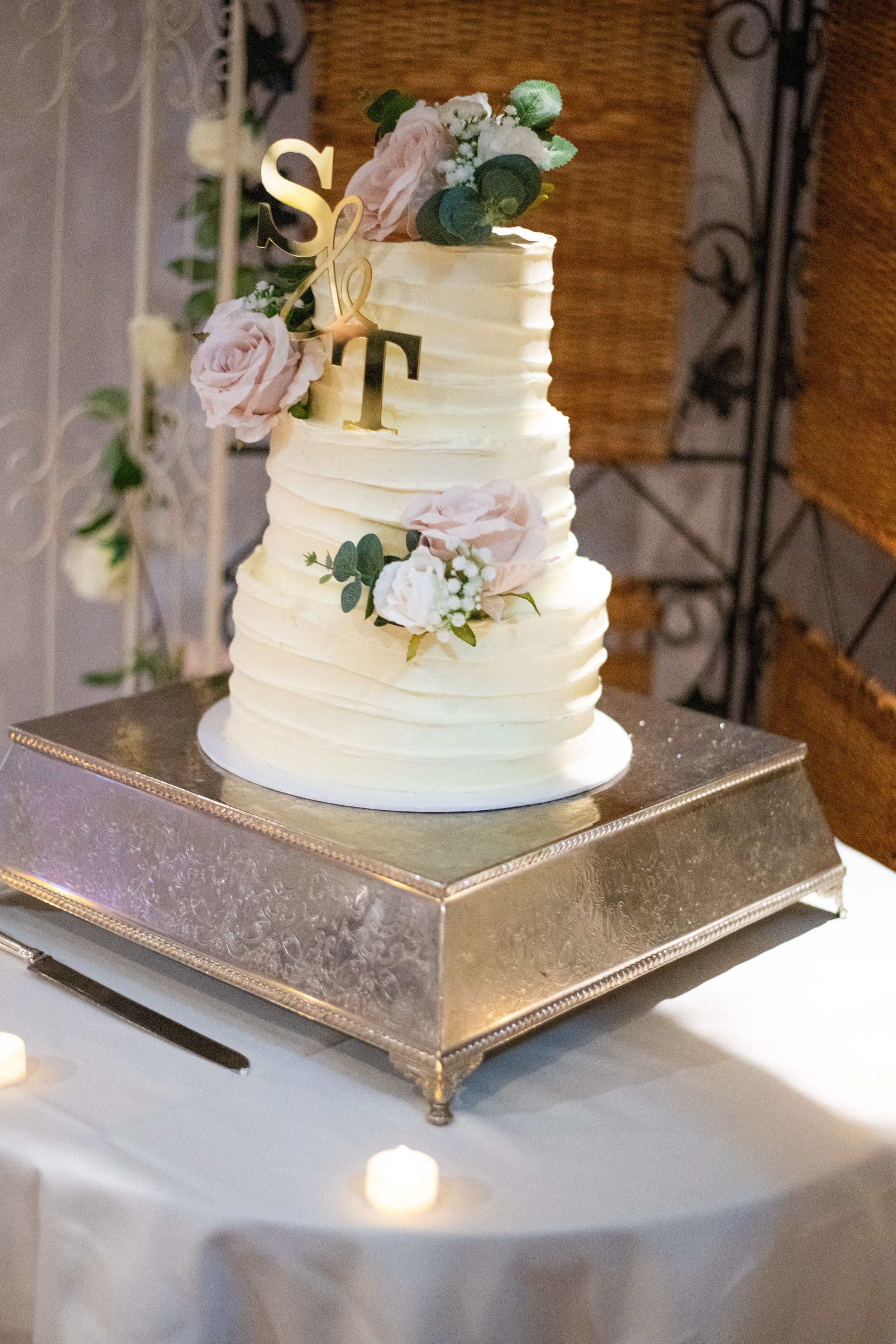Elegant white tiered wedding cake decorated with blush roses and monogram topper on silver stand.