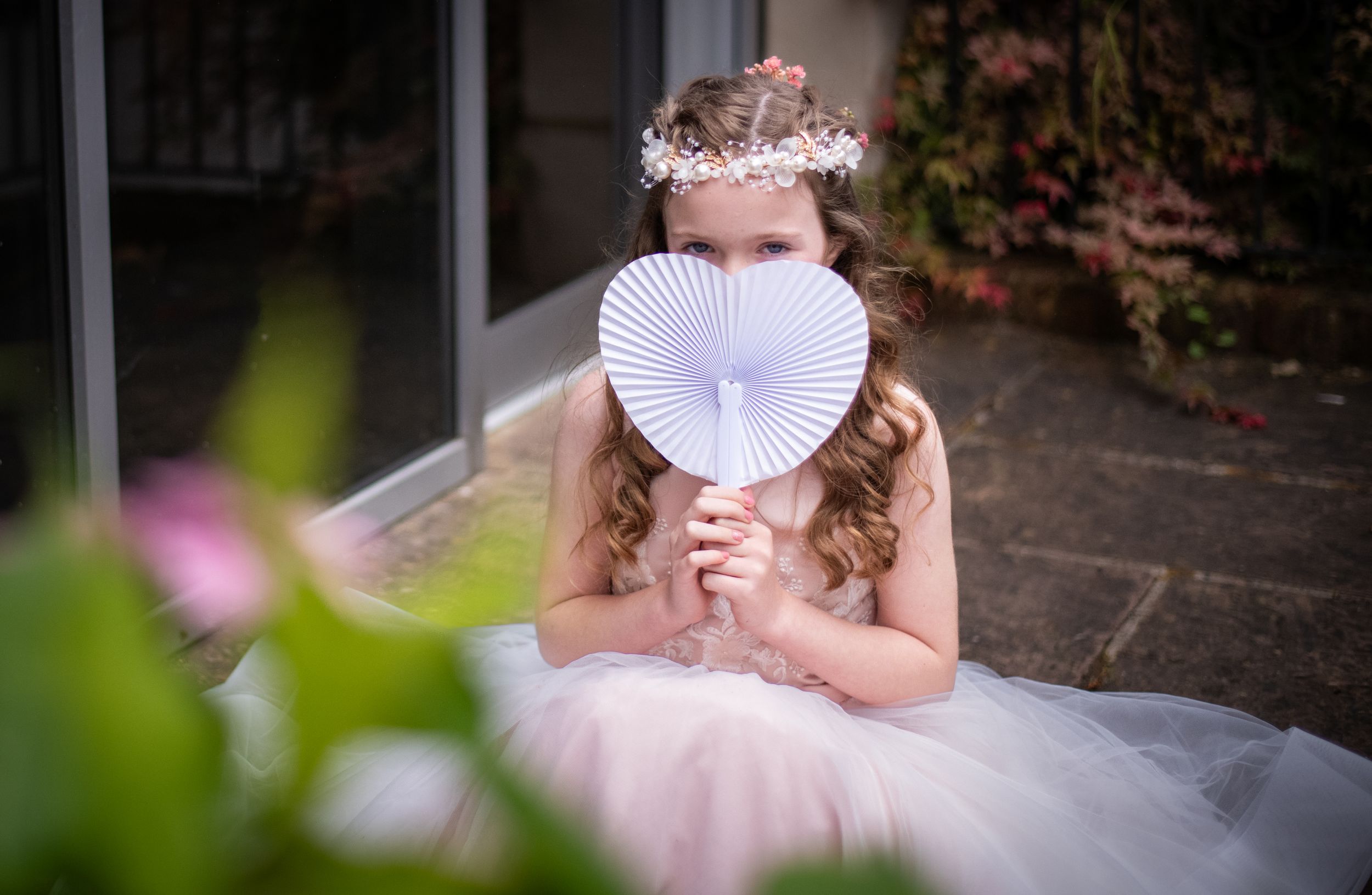 Dreamy portrait with white paper fan and pink dress amidst garden foliage.
