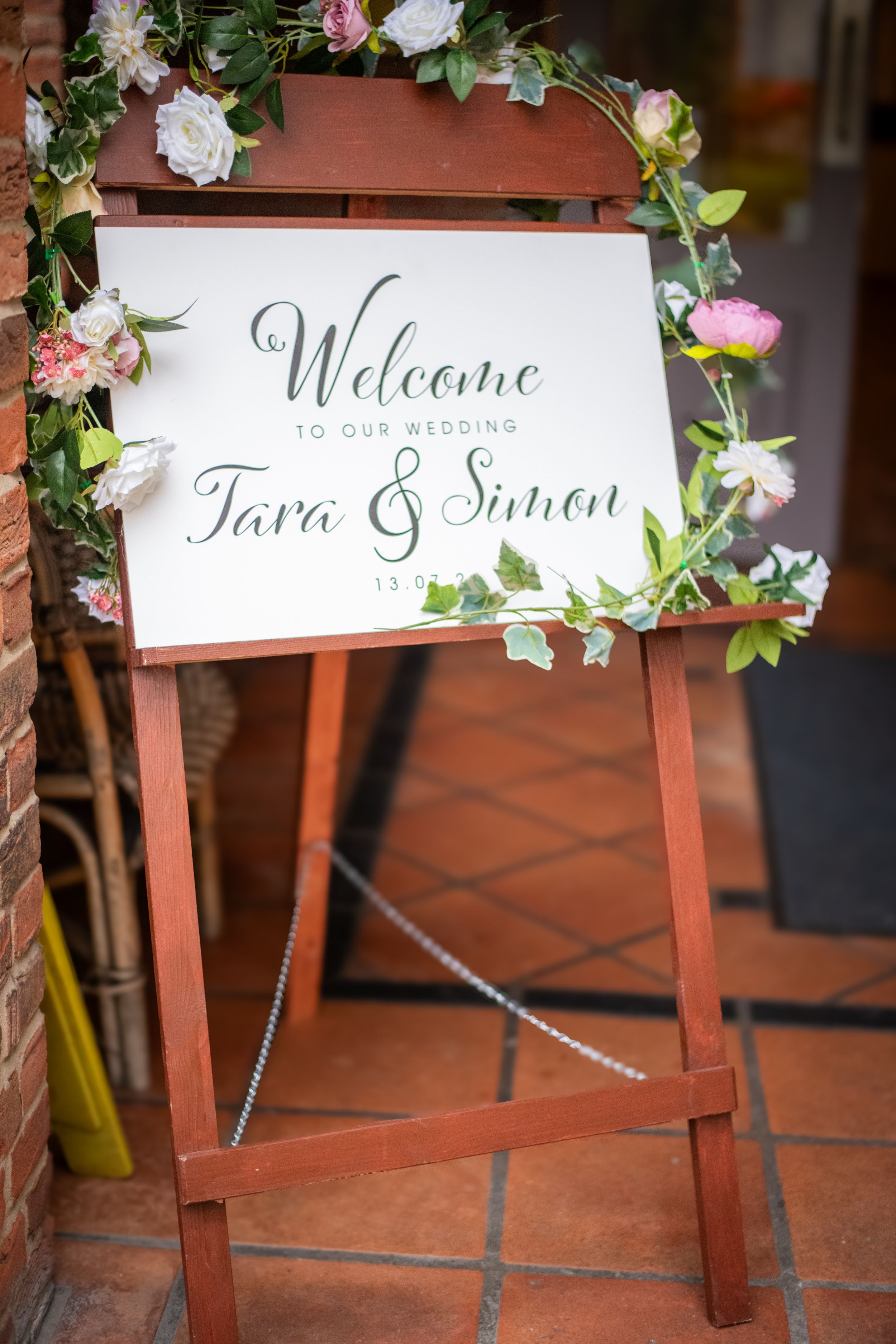 Wedding welcome sign decorated with pink roses and greenery on wooden easel.