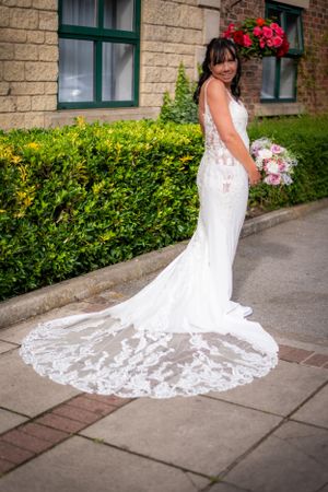 Bride in elegant lace wedding dress with long train poses outdoors against stone building with red flowers.