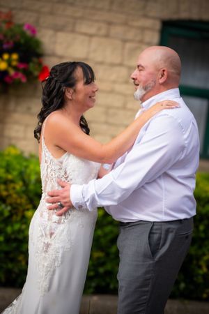 Wedding couple sharing joyful moment together outdoors in front of stone wall with colorful flowers.