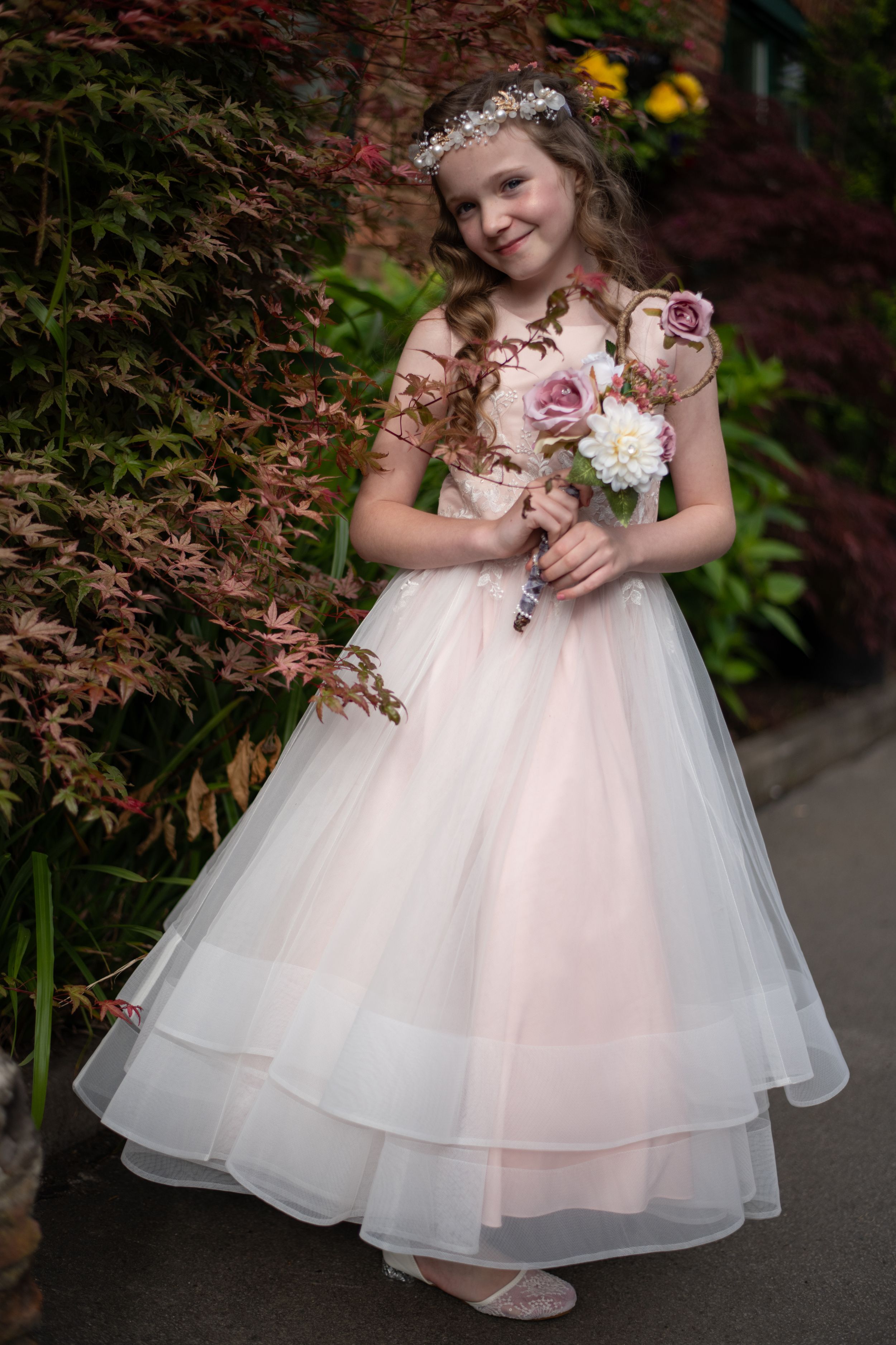 A princess-inspired flower girl in a flowing blush tulle gown holds a white flower in a garden setting.