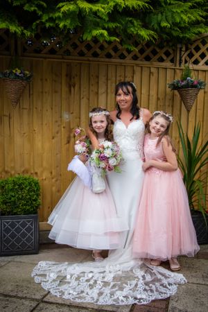 Wedding party portrait with flower girls in pink and white dresses against wooden fence backdrop.