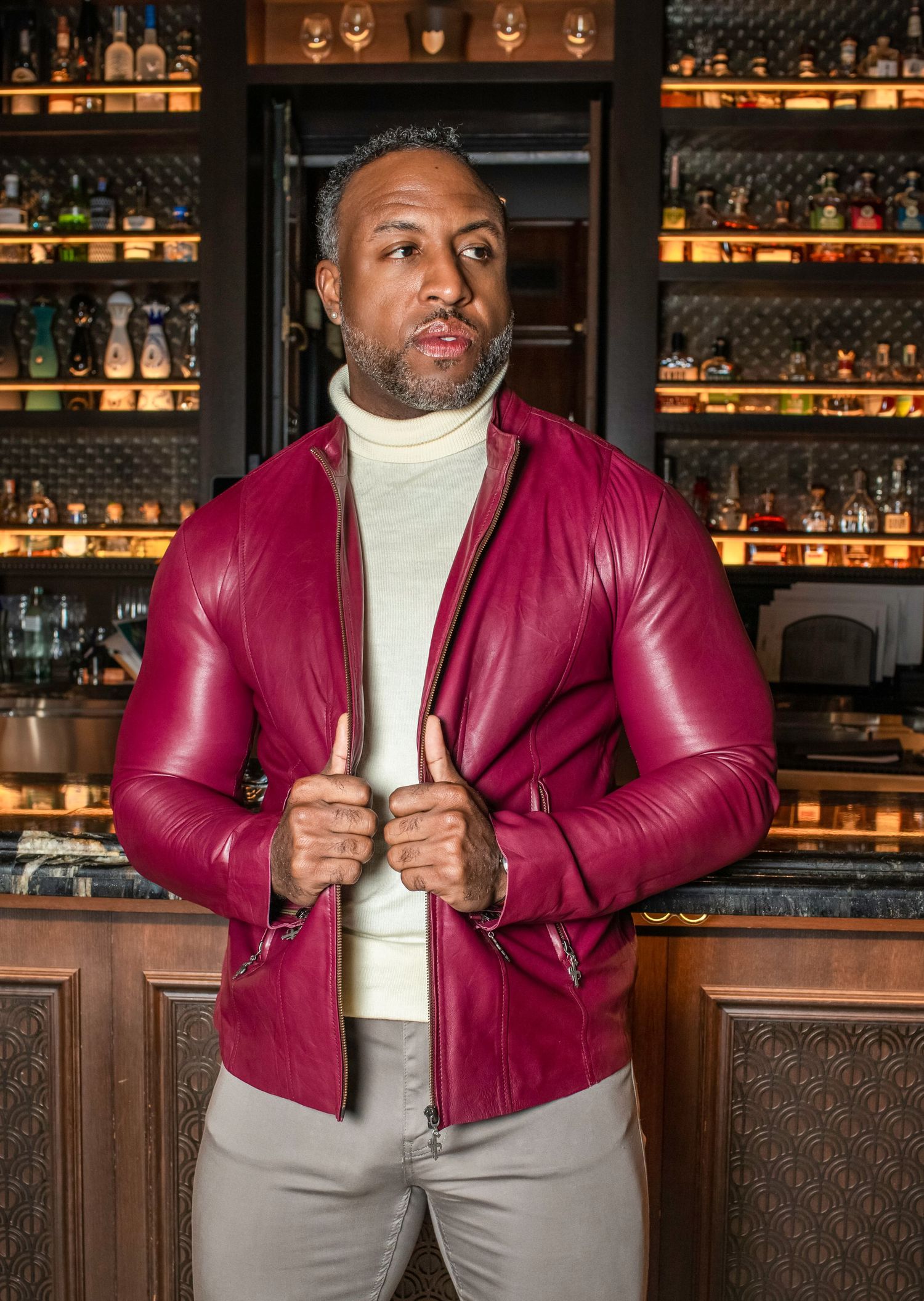 Stylish burgundy leather jacket paired with light gray pants and white turtleneck in upscale bar setting. Lifestyle, branding photography