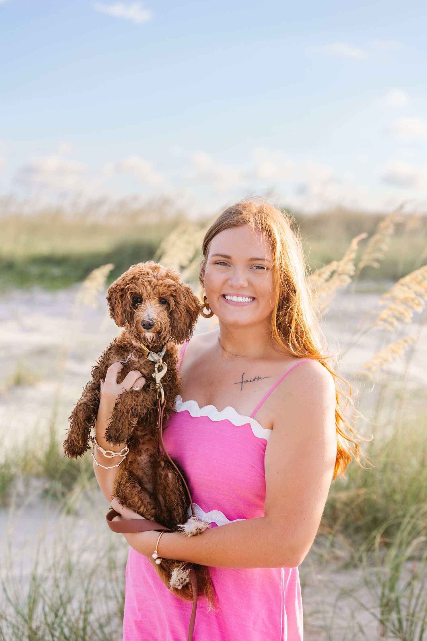 Person in pink dress holds brown goldendoodle puppy on beach with sea oats at sunset.