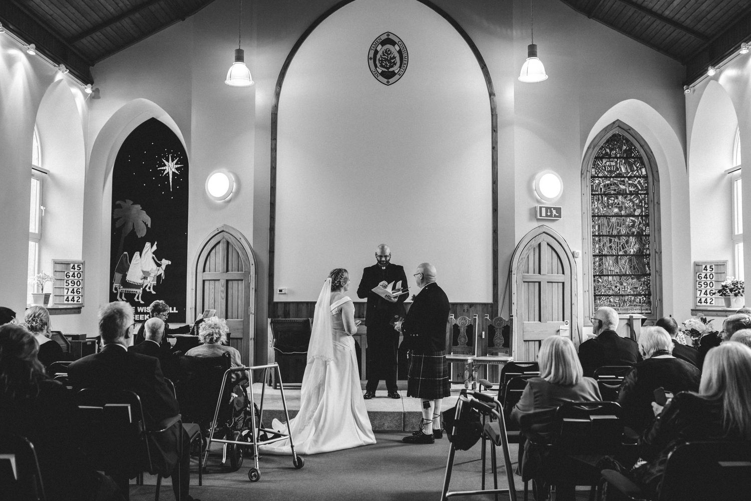 Black and white photograph of a traditional church wedding ceremony taking place in a vaulted chapel with stained glass windows.