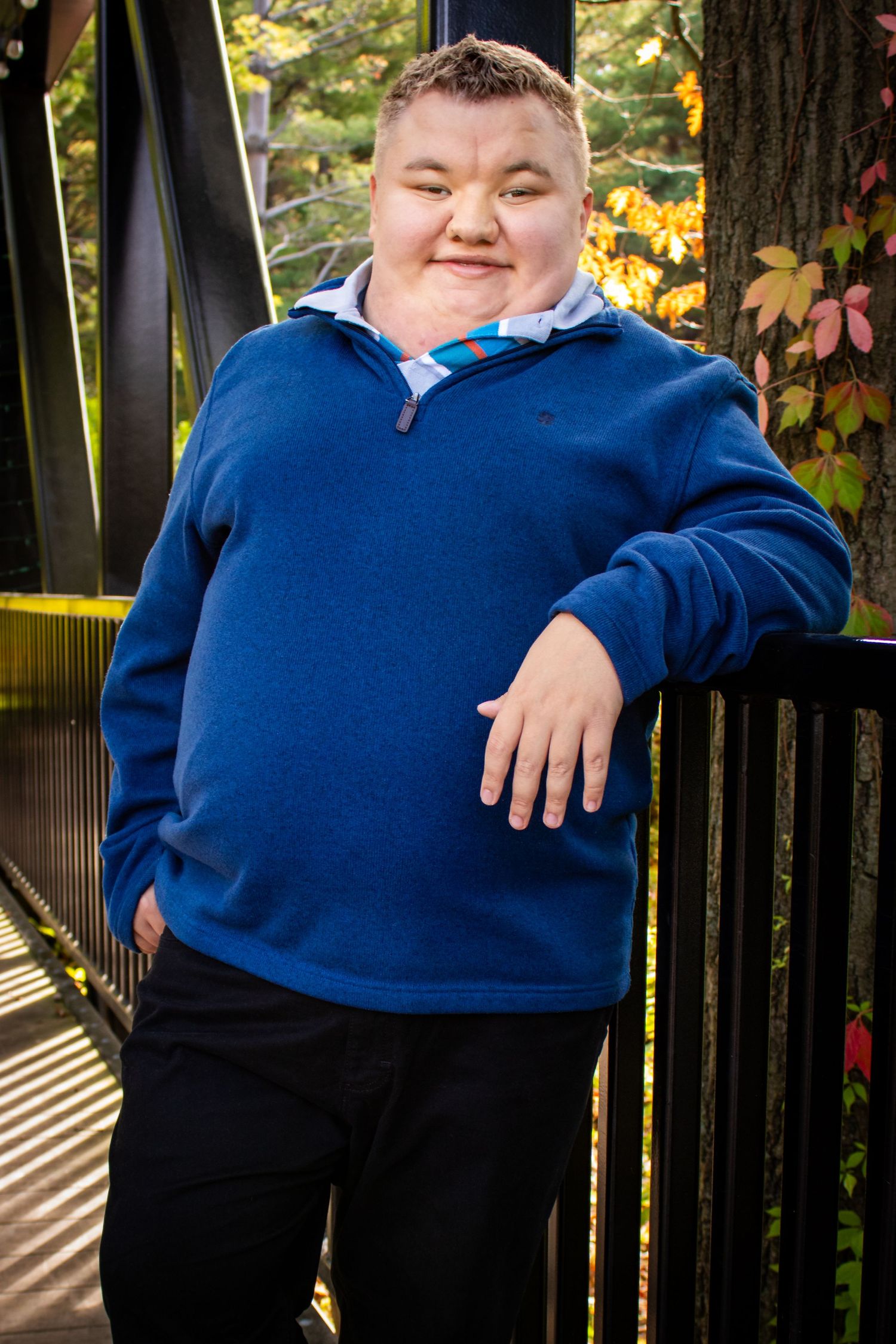 High school senior posing on a bridge with a confident smile, one hand in his pocket surrounded by fall foliage.