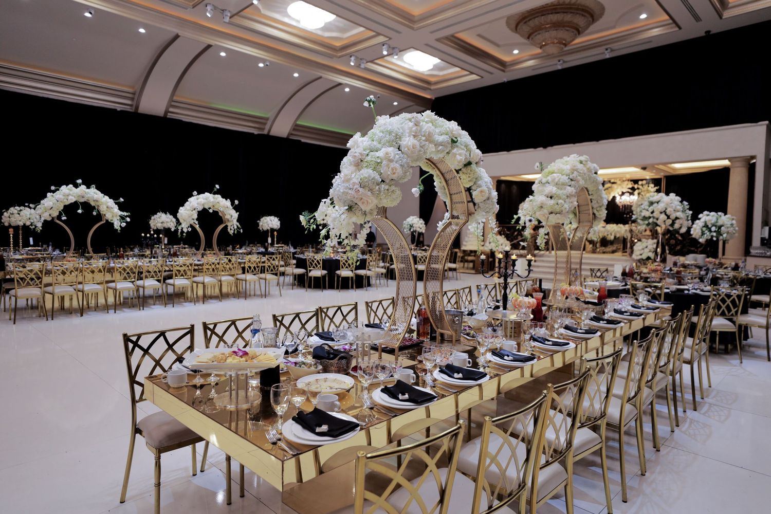 Elegant wedding reception setup with white floral arches and gold chairs along long banquet tables.