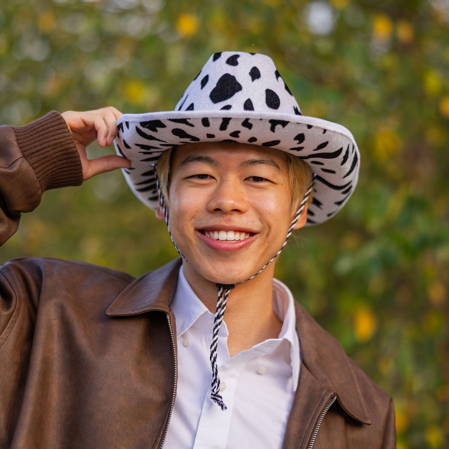 Alex Yuan wearing a cowboy hat and brown jacket smiling in front of greenery
