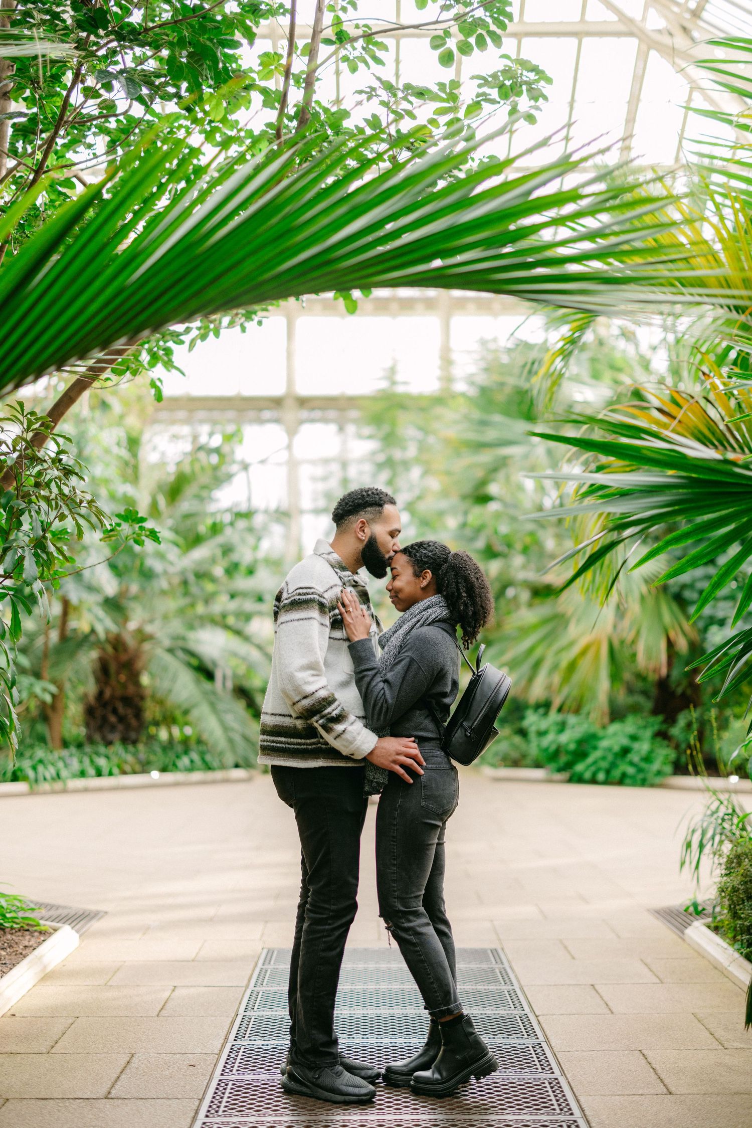 Romantic greenhouse engagement photo with tropical palm fronds framing a couple in casual attire.