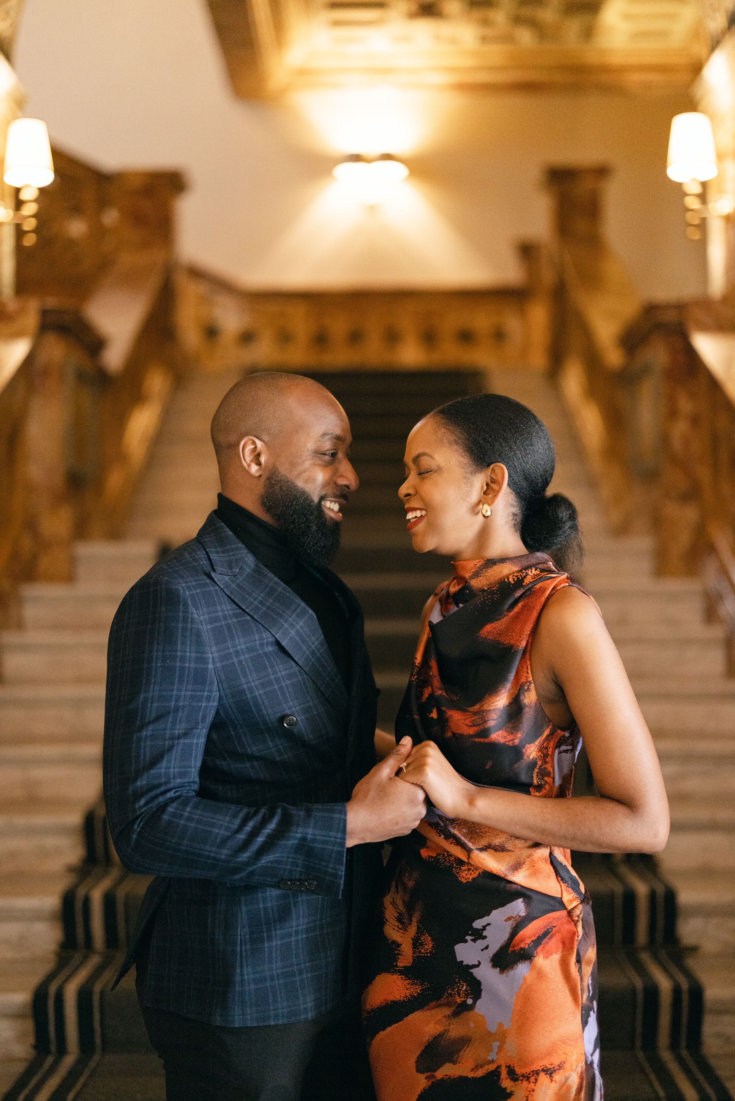A couple shares a warm embrace on an elegant staircase with warm lighting and luxurious decor.