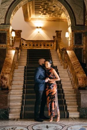 A romantic couple stands close together on an elegant marble staircase under decorative arches.