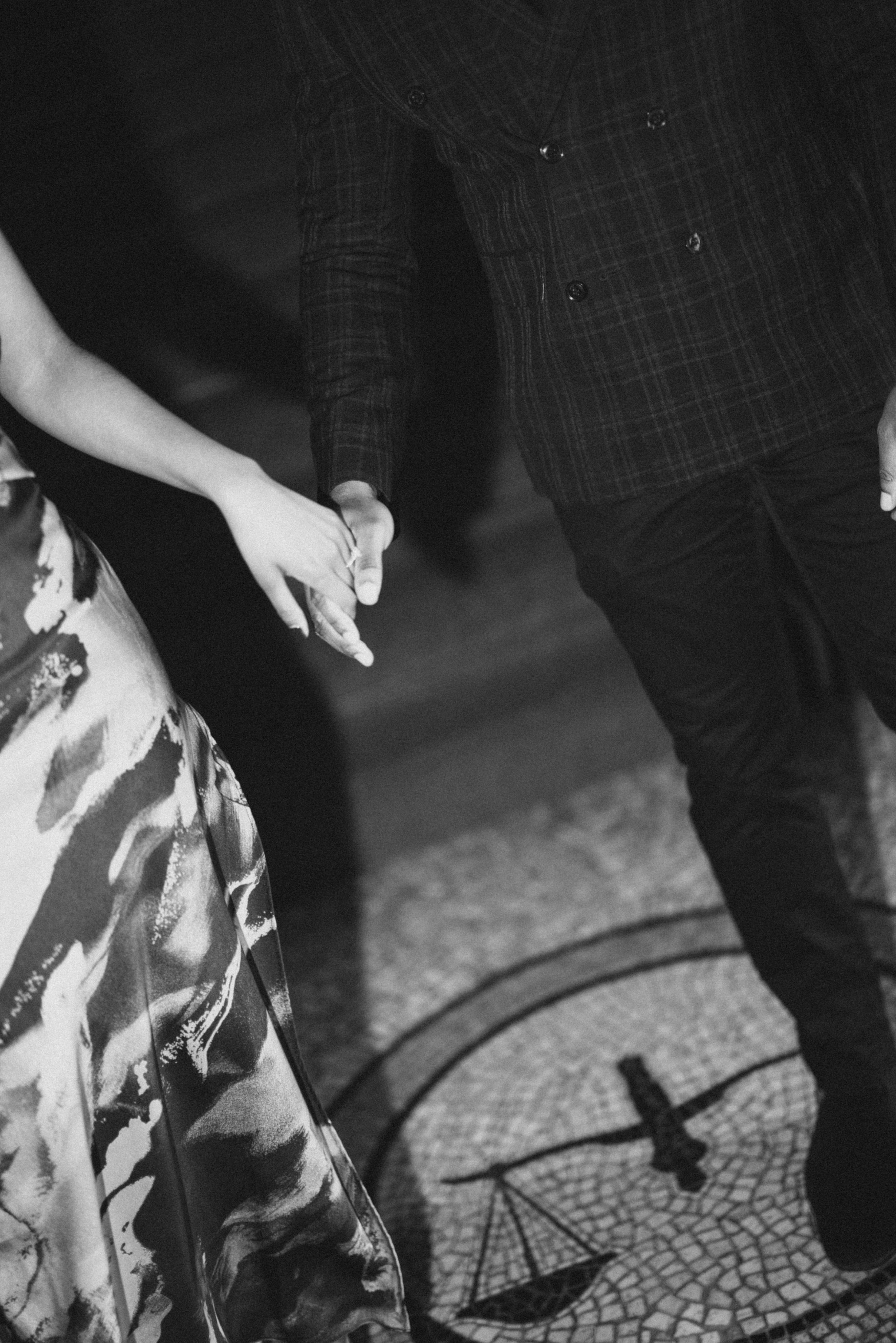 A black and white capture of joined hands against a patterned floor with dramatic shadows.
