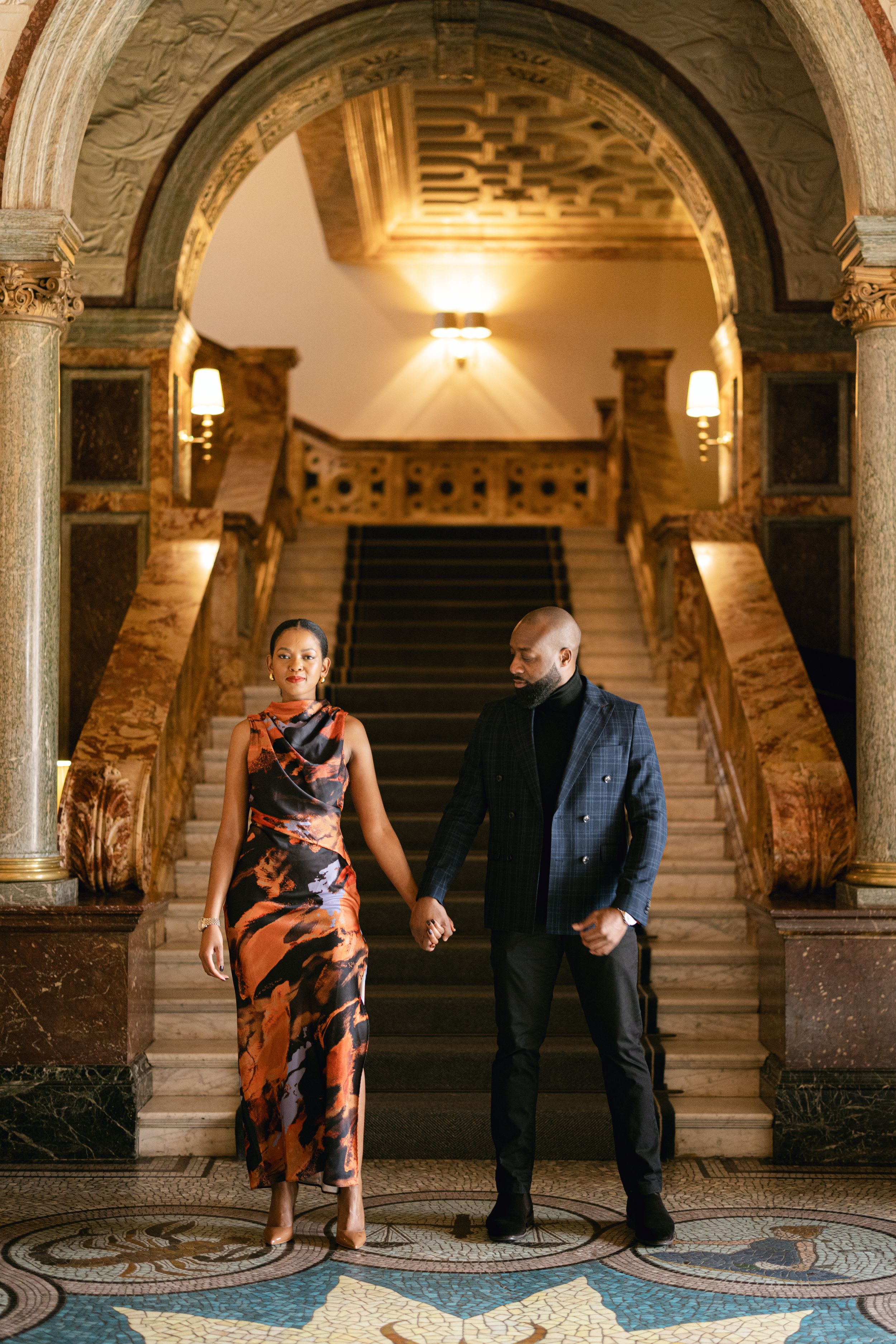 An elegant couple poses beneath an ornate archway with a grand staircase in the background.