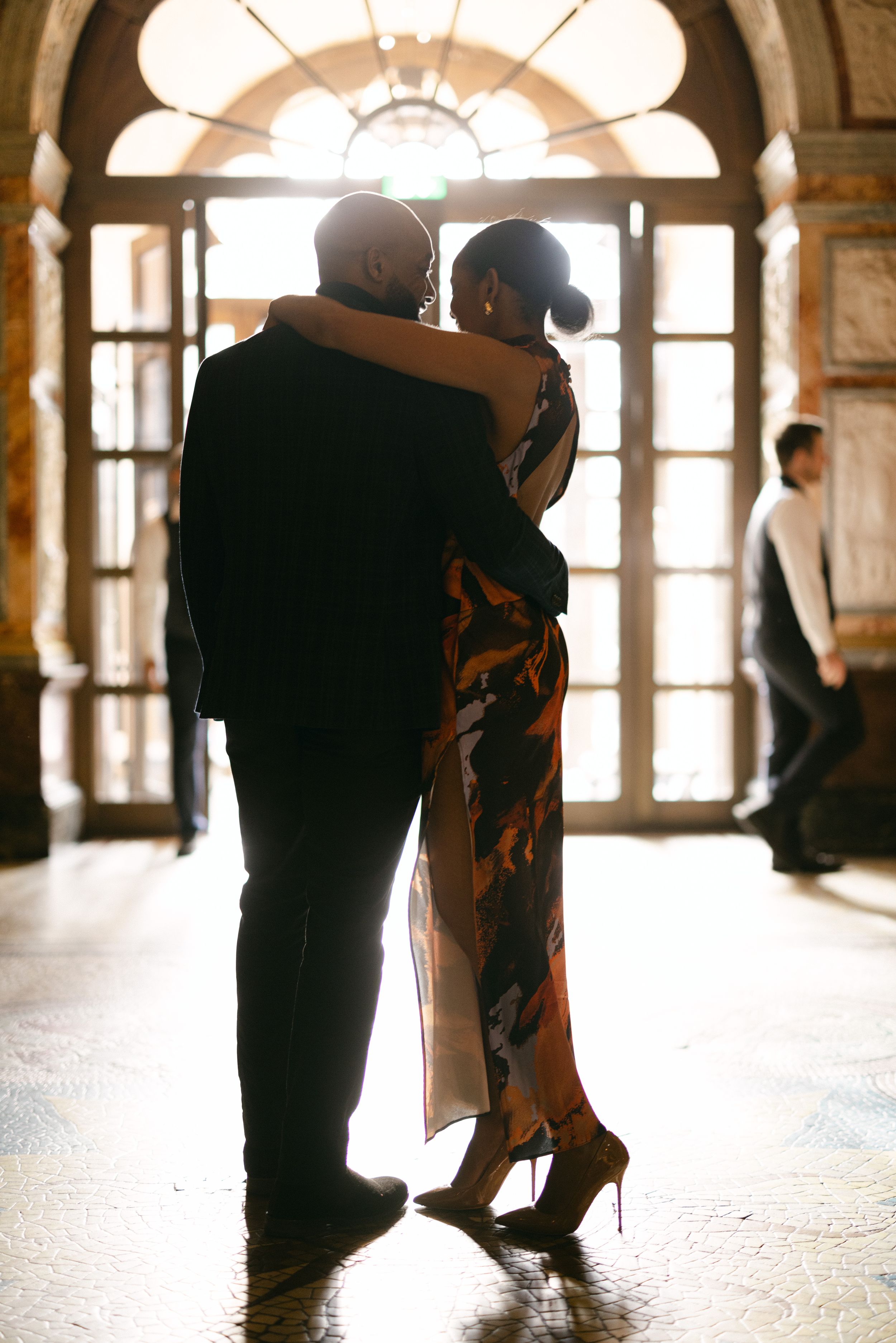 A silhouetted couple embraces in an ornate hallway with sunlit windows.