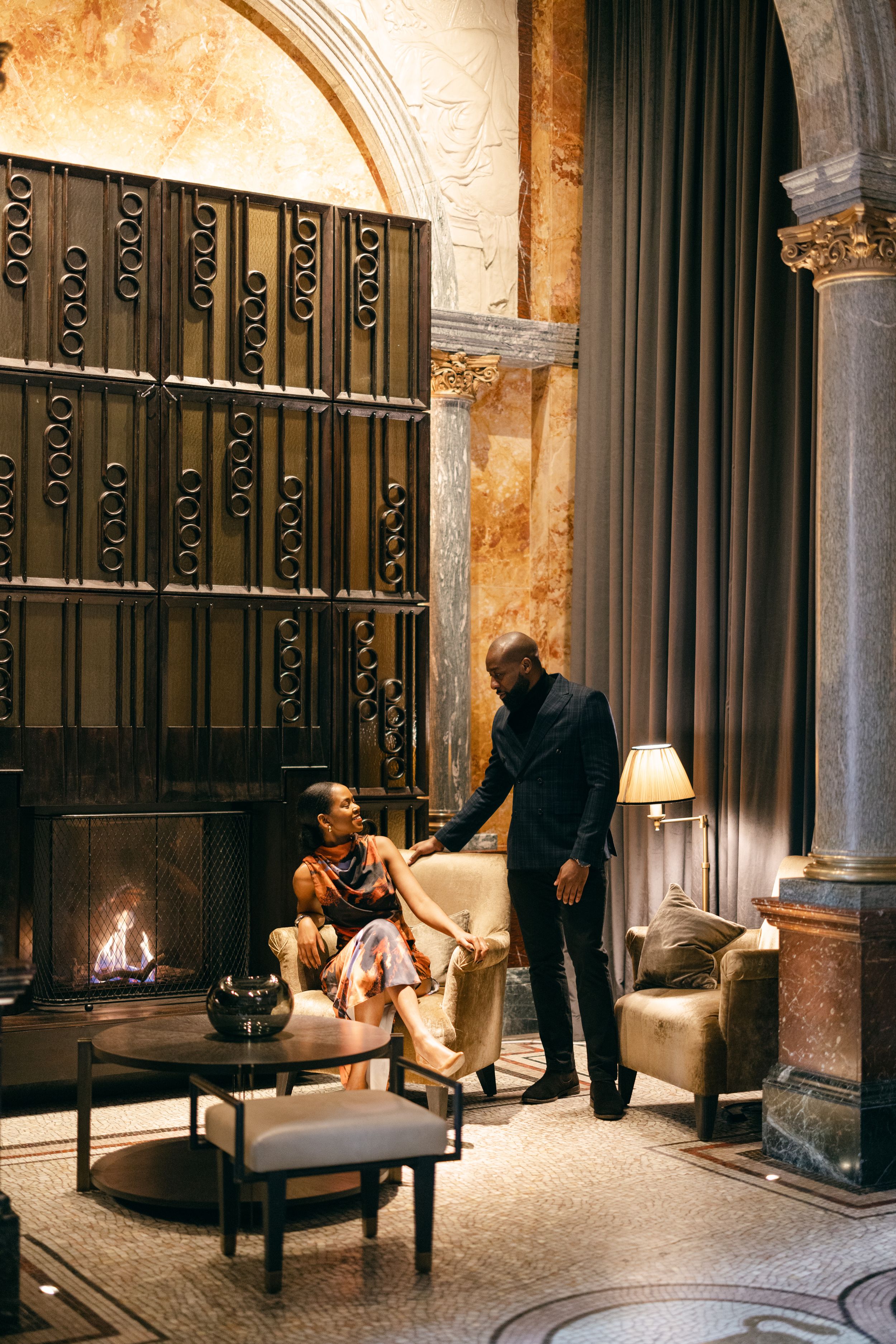 A couple shares a moment by a fireplace in an elegant room with ornate architectural details.