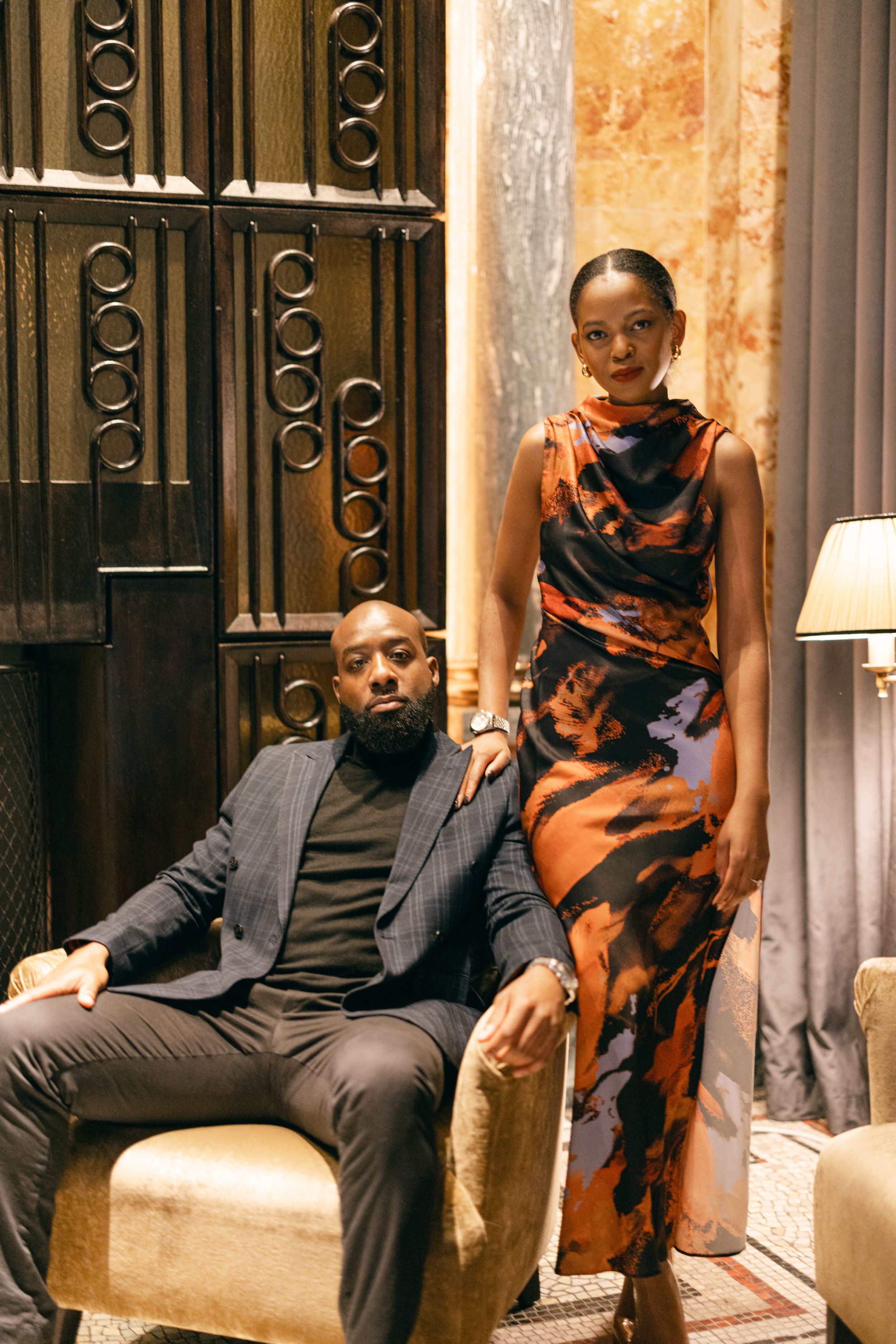 A stylish couple poses in a luxurious interior setting with decorative ironwork and warm lighting.